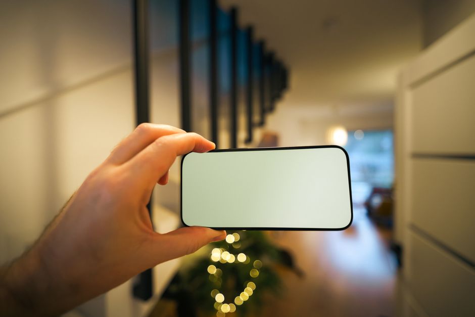 A hand holds a smartphone with a blank screen in a cozy, modern home interior with warm lighting.