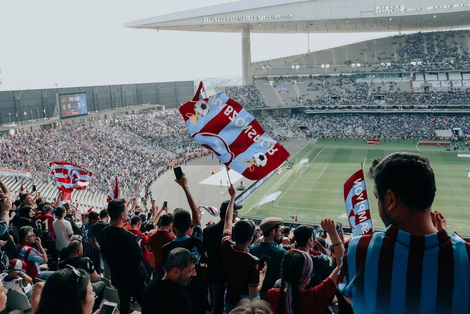 Vibrant crowd cheers at a football match in a large stadium, waving team flags.
