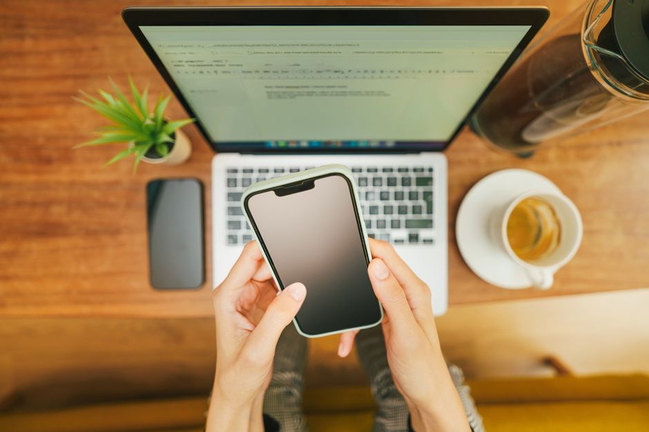 Hands holding a smartphone over a laptop with coffee in a cozy home office setup.